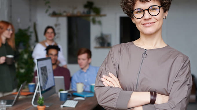 Mujer en primer plano mira de frente con expresin satisfecha mientras que detrs su equipo de trabajo realiza formacin online en la oficina